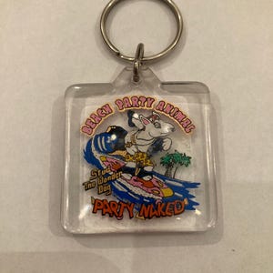 May include: A clear plastic keychain with a cartoon dog surfing on a pink and blue surfboard. The dog is wearing a yellow and brown shirt and a gold chain. The text "Beach Party Animal" and "Stud The Wonder Dog" are printed in pink. The text "Party Naked" is printed in red.