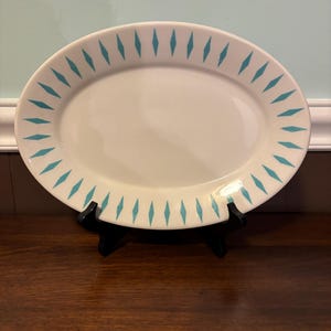 May include: An oval, off-white serving platter with a turquoise diamond pattern around the rim. The platter is displayed on a black stand, showcasing its design and shape. A vintage-style piece.