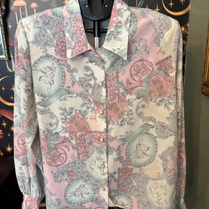 May include: A long-sleeved button-down blouse with a pastel pink, white, and gray baroque pattern. The design includes cherubs, musical instruments, and scrolls. The blouse is hanging on a black hanger.