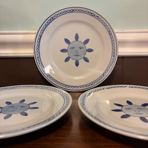 May include: Three white ceramic plates featuring a blue sun face illustration in the center and a blue chain-link pattern along the rim. The sun design includes a stylized face with rays extending outward, consistent across all three plates.