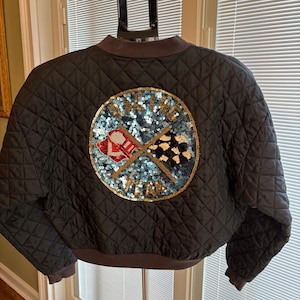 May include: Black quilted bomber jacket with a round sequined patch on the back. The patch features a red and white checkered flag, a gold and black checkered flag, and the word "Vitesse" in gold lettering.