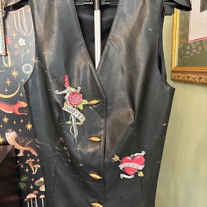 May include: Black leather vest with a V-neck and four gold-colored buttons. The vest features embroidered patches of a dagger and rose with the word "FOREVER", and a heart pierced by an arrow with the word "LOVE".