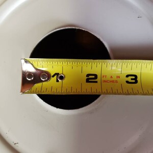K&ouml;nnte beinhalten: Ein gelbes Ma&szlig;band mit schwarzen Markierungen liegt auf einem wei&szlig;en runden Objekt. Das Band zeigt "1", "2 FT & IN INCHES" und "3" an.