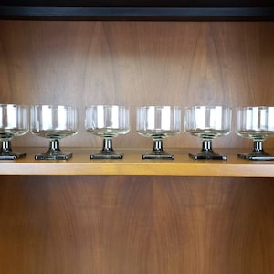 May include: Six vintage clear glass dessert bowls with a ribbed design. The bowls have a short stem and a square base. The bowls are arranged in a row on a wooden shelf, with a dark wooden background.