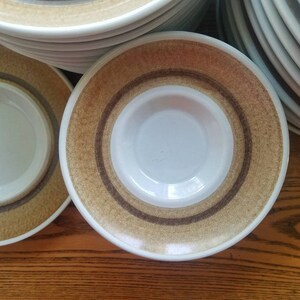 Puede incluir: Una pila de platillos de cerámica blanca con anillos concéntricos marrones y beige alrededor del centro. Los platillos están apilados sobre una superficie de madera.