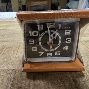 May include: A vintage Sunbeam alarm clock with a black face and white numerals. The clock is encased in a clear plastic wrap and set on a wooden base. The clock hands are red and silver.