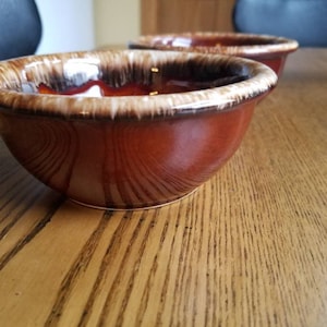 May include: Two brown ceramic bowls with a distinctive glaze. The bowls have a rounded shape with a lighter brown rim and a darker brown interior. The bowls are sitting on a wooden table.