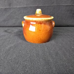 May include: A round, glazed ceramic pot with a lid, featuring a warm brown color palette. The pot has two small side handles and a central knob on the lid. The rim and lid edges show a lighter, yellowish-brown drip glaze effect on the surface.