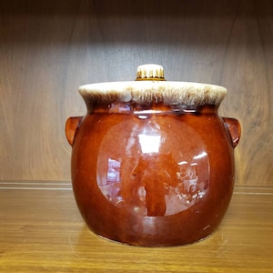 May include: A brown glazed ceramic crock with a lid. The crock has two handles and a white glaze around the top.