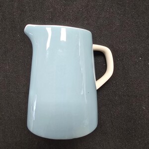 May include: A light blue ceramic pitcher with a white handle. The pitcher has a curved spout and a smooth, glossy finish. This kitchenware item is ideal for serving beverages.