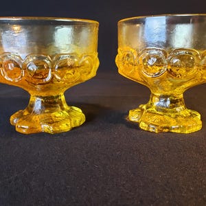 May include: Two vintage yellow glass dessert dishes. Each dish has a rounded bowl with a decorative pattern around the rim and sits on a pedestal base. The glass has a slight amber hue.