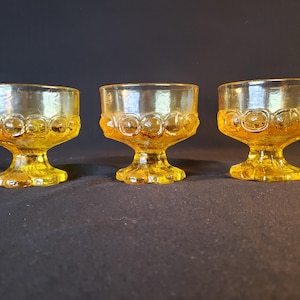May include: Three yellow glass dessert cups with a textured pattern. The cups have a short stem and a wide bowl.