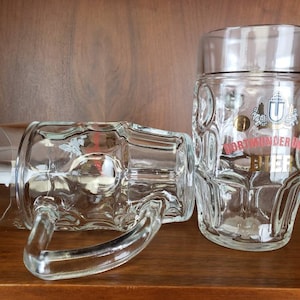 May include: Two clear glass beer steins with a raised design. The stein on the right has the text "Dortmunder Union Bier" in gold and red lettering.