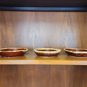 May include: Three brown and white glazed ceramic bowls are displayed on a wooden shelf. The bowls are oval shaped and have a brown glaze with white streaks.
