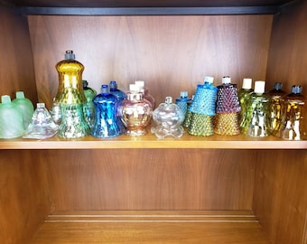 May include: A shelf filled with a variety of glass candle holders in different colors and styles. The candle holders are arranged in a row on the shelf, with some taller than others. The candle holders are made of clear, frosted, and colored glass.