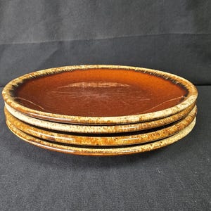 May include: A stack of three round ceramic plates featuring a glossy dark brown center that transitions into a lighter, mottled tan and cream rim. The plates have a smooth, reflective surface and a distinct color gradient from the middle to the edge.
