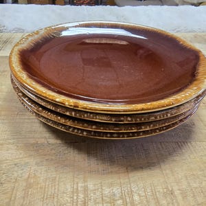 May include: A stack of four brown ceramic plates with a glossy finish. The plates have a wide, slightly raised rim with a lighter brown color. The plates are stacked on a wooden surface.
