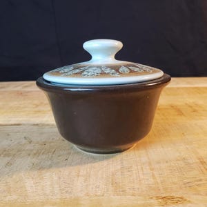 Puede incluir: Un cuenco de cerámica marrón con tapa. La tapa es de color azul claro con un asa blanca y un diseño decorativo en marrón y blanco. El cuenco está sobre una superficie de madera.