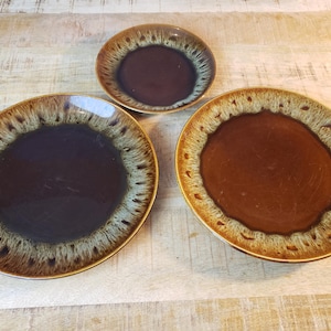 May include: Three ceramic plates with a brown center and a speckled, light brown and cream-colored rim. The plates vary in size, with a small, medium, and large plate. The plates are on a light brown wooden surface.