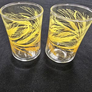 May include: Two clear drinking glasses with a yellow wheat stalk design. The glasses have a slightly tapered shape and are made of clear glass. The yellow design is a repeating pattern of wheat stalks.