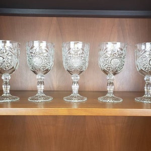 May include: Five clear glass goblets with a cut glass design sit on a wooden shelf. The goblets are all the same size and shape, and they have a round base and a tall, slender stem.