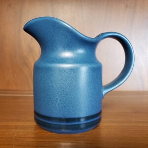 May include: A blue ceramic creamer with a black band around the bottom. The creamer has a rounded body and a handle.