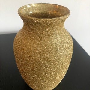 May include: A gold-colored vase with a textured, glittery surface. The vase has a rounded body and a flared rim. The vase is likely made of glass or a similar material. The overall shape is reminiscent of a classic urn.