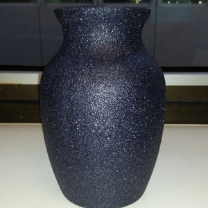 May include: A dark blue, glitter-covered vase with a wide base and a flared rim. The vase has a textured surface and a classic, elegant shape. The vase is likely made of ceramic or glass.