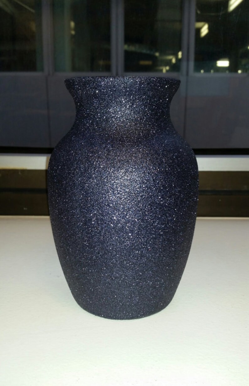 May include: A dark blue, glitter-covered vase with a wide base and a flared rim. The vase has a textured surface and a classic, elegant shape. The vase is likely made of ceramic or glass.