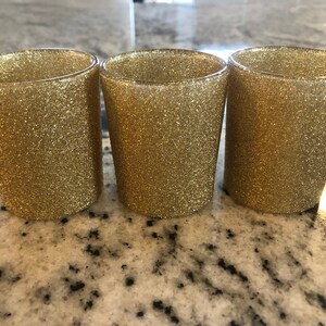 May include: Three gold glitter shot glasses. The glasses are cylindrical with a slightly flared top. The surface of each glass is covered in gold glitter, giving them a sparkling appearance. They are arranged in a row on a light-colored surface.