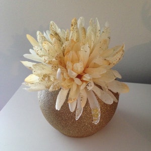 May include: A decorative floral arrangement featuring a large, multi-layered flower in shades of cream and peach, dusted with gold glitter. The flower sits in a round, gold glitter-covered vase, creating a glamorous centerpiece. The arrangement is ideal for home decor.