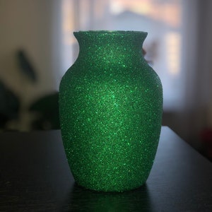 May include: A green glitter-covered vase with a flared top. The vase has a rounded body and a narrow neck. The glitter gives the vase a textured appearance, reflecting light. The vase is sitting on a dark surface.