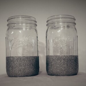 May include: Two clear glass mason jars with silver glitter applied to the bottom half. The jars have a classic design with the word "Ball" embossed on the front. The jars are likely used for home decor or storage.