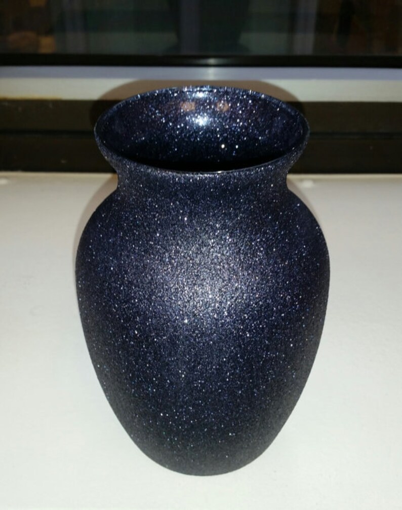 May include: A navy blue vase with a glittery, textured surface. The vase has a rounded body and a flared rim. The glitter catches the light, creating a sparkling effect. This decorative vase is suitable for displaying flowers or as a standalone accent piece.