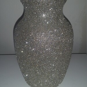 May include: A silver glitter vase with a wide body and a narrow neck. The vase has a classic urn shape and is covered in sparkling glitter, reflecting light. The vase is a decorative item.