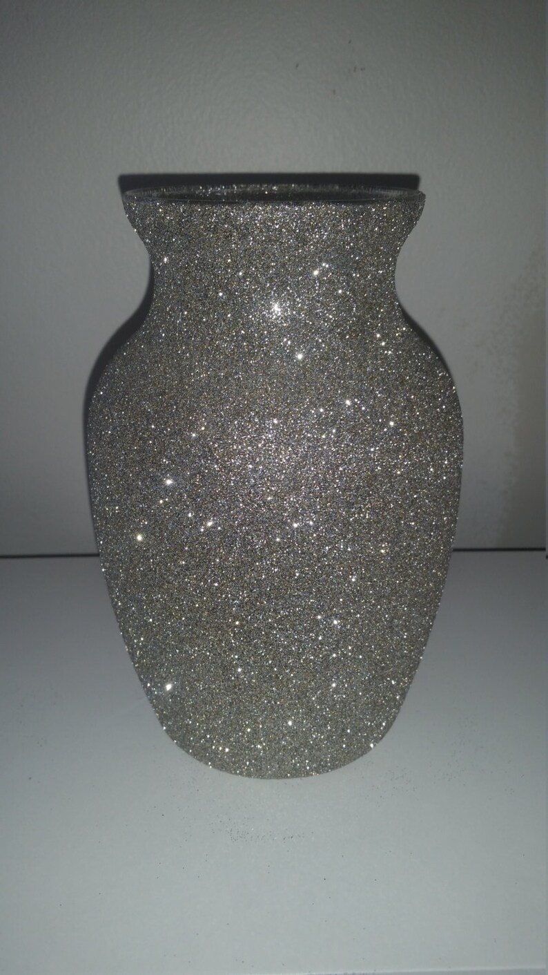 May include: A silver glitter vase with a wide body and a narrow neck. The vase has a classic urn shape and is covered in sparkling glitter, reflecting light. The vase is a decorative item.
