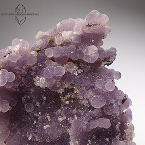 Puede incluir: Un racimo de ágata púrpura con una textura lisa y redondeada. La ágata es de color púrpura claro y está cubierta de pequeños cristales blancos.