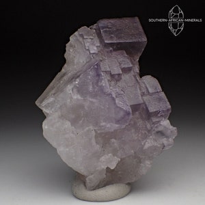 May include: A cluster of purple fluorite crystals with a cubic habit. The crystals are translucent and have a glassy luster. The fluorite is mounted on a gray base.