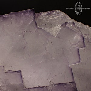May include: Close-up of a crystalline mineral with a layered, geometric structure. The mineral has a pale purple hue with darker purple accents, and a rough, textured surface. The image includes the text "SOUTHERN-AFRICAN-MINERALS".