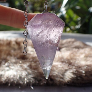 Puede incluir: Un péndulo de cristal de amatista púrpura con una cadena de plata. El péndulo está facetado y tiene una punta puntiaguda.