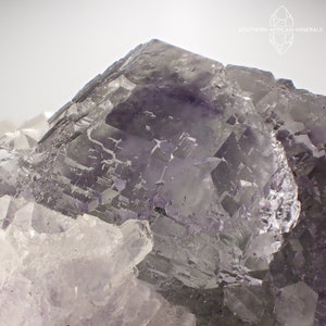 May include: Close-up of a fluorite crystal formation. The crystal features a mix of purple and clear sections, with a geometric, cube-like structure. The image is well-lit, highlighting the mineral's facets and textures. The Southern-African-Minerals logo is in the top right corner.