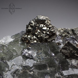 Peut inclure: Un groupe de cristaux de fluorine gris-vert avec un grand cristal de pyrite gris métallique au sommet. Le cristal de pyrite est composé de nombreux petits cristaux regroupés.