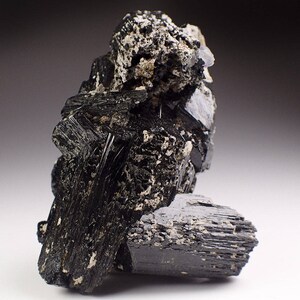 May include: A cluster of black schorl tourmaline crystals with a rough, textured surface. The crystals are arranged in a random, natural formation.