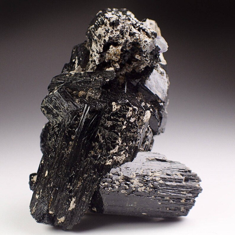 May include: A cluster of black schorl tourmaline crystals with a rough, textured surface. The crystals are arranged in a random, natural formation.