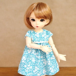 Peut inclure: Une poupée portant une robe bleue à imprimé floral blanc. La robe est ornée d'un ruban blanc à la taille. La poupée a les cheveux bruns et les yeux bleus.