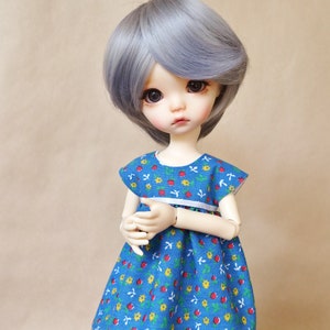 Puede incluir: Muñeca con cabello gris y un vestido azul. El vestido tiene una cintura blanca y un estampado de fresas rojas, flores amarillas y hojas blancas. La muñeca está sobre una superficie de madera.