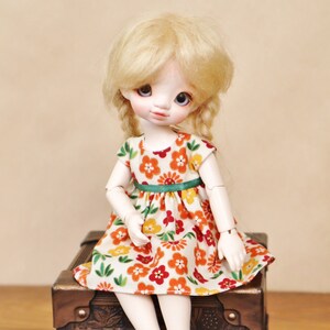 May include: A doll with blonde hair in pigtails wearing a floral print dress with red, orange, and yellow flowers. The doll is sitting on a small, dark brown wooden chest with metal corner accents.