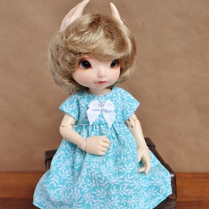 Può includere: Una bambola con capelli biondi e occhi azzurri che indossa un vestito azzurro chiaro con un motivo a foglie bianche e un fiocco bianco. La bambola è seduta su uno scrigno di legno.