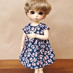 Peut inclure: Une poupée aux cheveux courts et bruns clairs portant une robe bleue à motifs floraux blancs et rouges. La poupée a le teint clair et se tient debout, les bras croisés. Les pieds de la poupée sont nus.