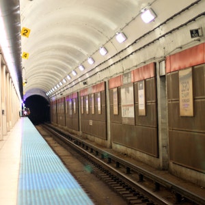 May include: A view down a subway platform with a blue tiled floor and a train track in the distance. The platform is lined with white pillars and posters on the wall. The posters advertise a local business called "Grand Central", and a sign reads "10N".